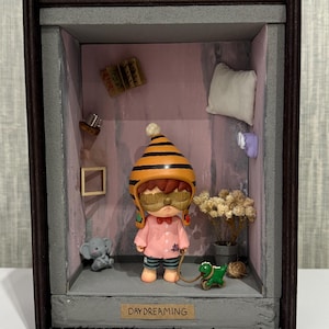 May include: A miniature diorama featuring a figure in a pink shirt and striped helmet, holding a leash attached to a green dinosaur toy. The scene includes a small elephant, dried flowers, and the text "DAYDREAMING". The diorama is enclosed in a dark wooden frame.