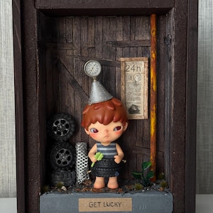 Puede incluir: Un diorama con una muñeca de pelo castaño y un gorro de fiesta, con una camisa a rayas y una falda de neumáticos. La escena incluye neumáticos en miniatura, un medidor y un letrero que dice "GET LUCKY". El fondo es una puerta de madera.