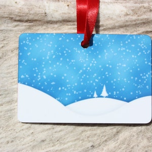 May include: Rectangular Christmas ornament with a winter scene. The ornament features a blue sky with falling snow and a white landscape with two trees. A red ribbon is attached for hanging.