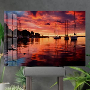 Könnte beinhalten: Ein lebendiger Sonnenuntergang über einem ruhigen Hafen, mit mehreren Segelbooten, die sich vor dem feurigen Himmel abzeichnen. Das Wasser spiegelt die orangefarbenen und roten Farbtöne der Wolken wider und erzeugt eine friedliche Szene. Ein dekoratives Stück für jeden Raum.