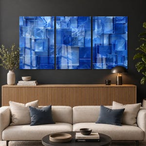 Puede incluir: Un tr&iacute;ptico de paneles de arte abstracto con un dise&ntilde;o geom&eacute;trico de bloques de vidrio azules. La obra de arte se exhibe sobre un mueble de madera, con un sof&aacute; beige y cojines azul oscuro en frente. Una l&aacute;mpara negra y objetos decorativos completan la escena.