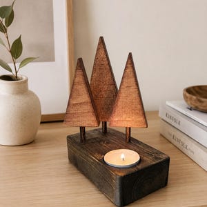 May include: A dark brown wooden candle holder featuring three triangular tree-shaped cutouts of varying heights attached to a rectangular base. A circular cutout in the base holds a small, lit tea light candle, casting a warm glow on the wood.
