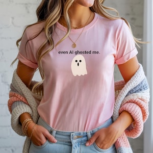 May include: A pale pink t-shirt featuring the phrase "even AI ghosted me." and a cartoon ghost illustration. The ghost is white with black eyes and a black mouth. The shirt has short sleeves and a round neck.