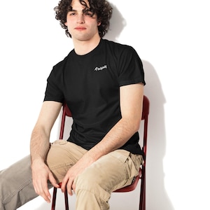 Puede incluir: Camiseta negra de cuello redondo con la palabra "behind!" impresa en blanco en el pecho. La camiseta se usa con pantalones cargo color caqui. La persona está sentada en una silla roja sobre un fondo blanco.