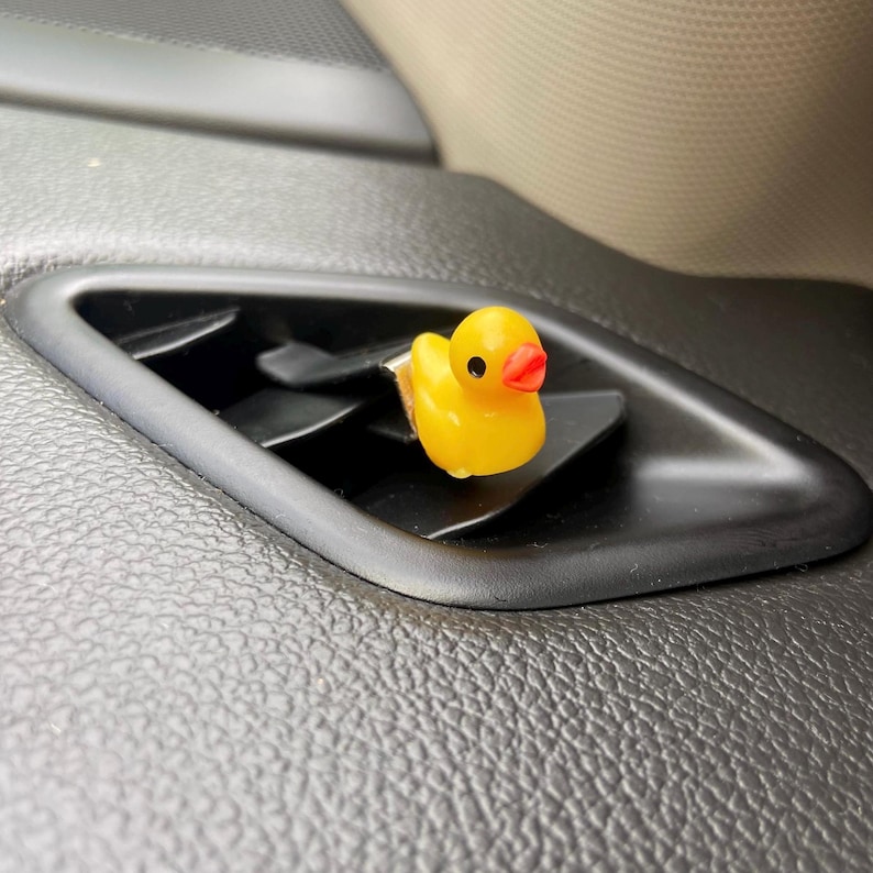 May include: A yellow rubber ducky air freshener hanging from the air vent of a car.