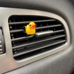 May include: A yellow rubber ducky car air freshener hanging from a car's air vent.