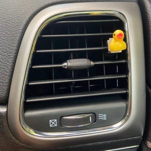 May include: A yellow rubber duck car air freshener hanging from a car's air vent. The air vent has a black plastic knob and a silver trim.