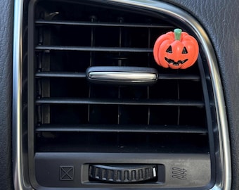 Puede incluir: Un pequeño clip ambientador con forma de calabaza naranja, con un tallo verde y rasgos faciales negros tallados, sujeto a las lamas horizontales de una rejilla de ventilación negra de un vehículo con un borde metálico plateado.