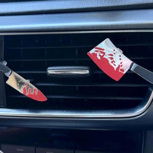 May include: Two car air vent clips shaped like a kitchen knife and a meat cleaver. Both items feature a metallic silver blade finish with realistic red blood splatter patterns and black handles, securely attached to the horizontal slats of a car vent.