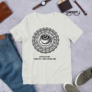 May include: A white t-shirt featuring a black graphic of a coffee cup within a chain ring. The text "Fueled by Coffee and Chain Oil" is printed beneath the graphic.