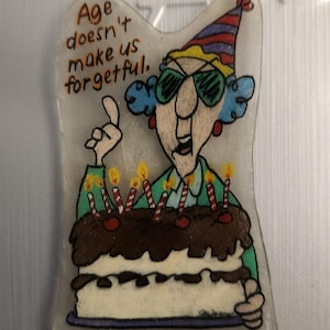 Può includere: Un magnete rettangolare trasparente con un'illustrazione a cartoni animati di una donna con un cappello da festa e occhiali da sole, che tiene in mano una torta di compleanno con candele accese. Il testo recita: "L'età non ci rende smemorati."