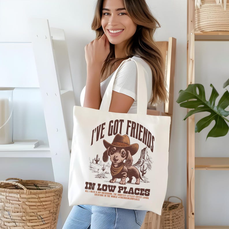 May include: Cream-colored tote bag featuring a cartoon dachshund wearing a cowboy hat and bandana, with the text "I'VE GOT FRIENDS IN LOW PLACES". The design includes desert scenery and additional text at the bottom.