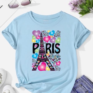 May include: Light blue t-shirt featuring a colorful graphic of the Eiffel Tower with the word "PARIS" in bold black letters. The design includes hearts and leopard print accents, creating a vibrant and eye-catching look.