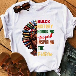 May include: White t-shirt with a colorful illustration of a woman with books in her hair. The text "BLACK HISTORY HONORING The past INSPIRING THE FUTURE" is printed on the shirt.