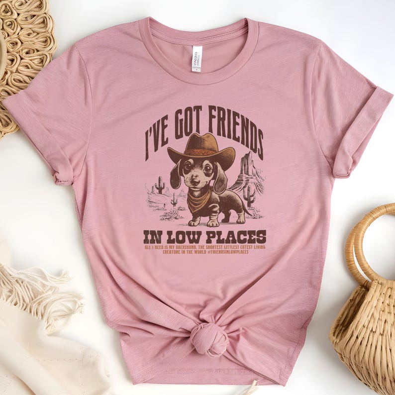May include: Dusty rose t-shirt featuring a dachshund wearing a cowboy hat and bandana, with the text "I'VE GOT FRIENDS IN LOW PLACES". The graphic includes desert scenery and the phrase "ALL I NEED IS MY DACHSHUND, THE SHORTEST CUTEST LIVING CREATURE IN THE WORLD".