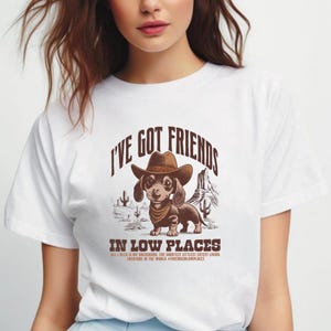 May include: White t-shirt featuring a dachshund wearing a cowboy hat and bandana, with the text "I'VE GOT FRIENDS IN LOW PLACES." The graphic includes desert scenery and additional text: "ALL I NEED IS MY DACHSHUND, THE SHORTEST LITTLEST CUTEST LIVING CREATURE IN THE WORLD #FRIENDSINLOWPLACES."
