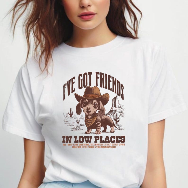 May include: White t-shirt featuring a dachshund wearing a cowboy hat and bandana, with the text "I'VE GOT FRIENDS IN LOW PLACES." The graphic includes desert scenery and additional text: "ALL I NEED IS MY DACHSHUND, THE SHORTEST LITTLEST CUTEST LIVING CREATURE IN THE WORLD #FRIENDSINLOWPLACES."