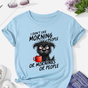 May include: Pale blue t-shirt with rolled sleeves. It features a graphic of a sleepy black dog holding a red mug, with the text "I DON'T LIKE MORNING PEOPLE OR MORNINGS OR PEOPLE."