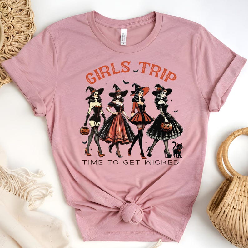 May include: A dusty rose t-shirt featuring a graphic of four women dressed as witches, with the words "GIRLS TRIP" and "TIME TO GET WICKED" in orange. The witches are holding pumpkins and a black cat is present.