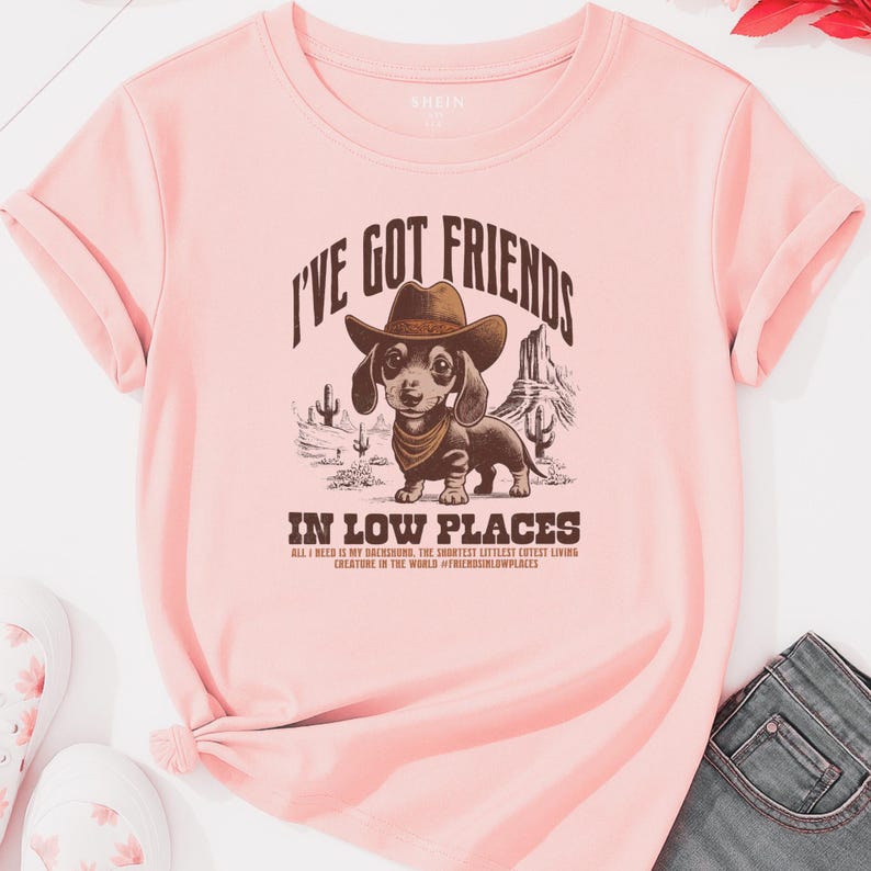 May include: A light pink t-shirt with rolled sleeves featuring a graphic of a dachshund wearing a cowboy hat and bandana. The text reads "I'VE GOT FRIENDS IN LOW PLACES" with additional text below.