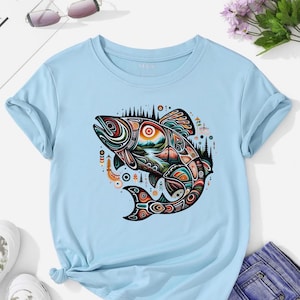 May include: Pale blue t-shirt with a vibrant, patterned fish graphic. The design includes a mountain and forest scene, with colours such as red, orange, green, and black. The top has a crew neck and short sleeves, suitable for casual wear.