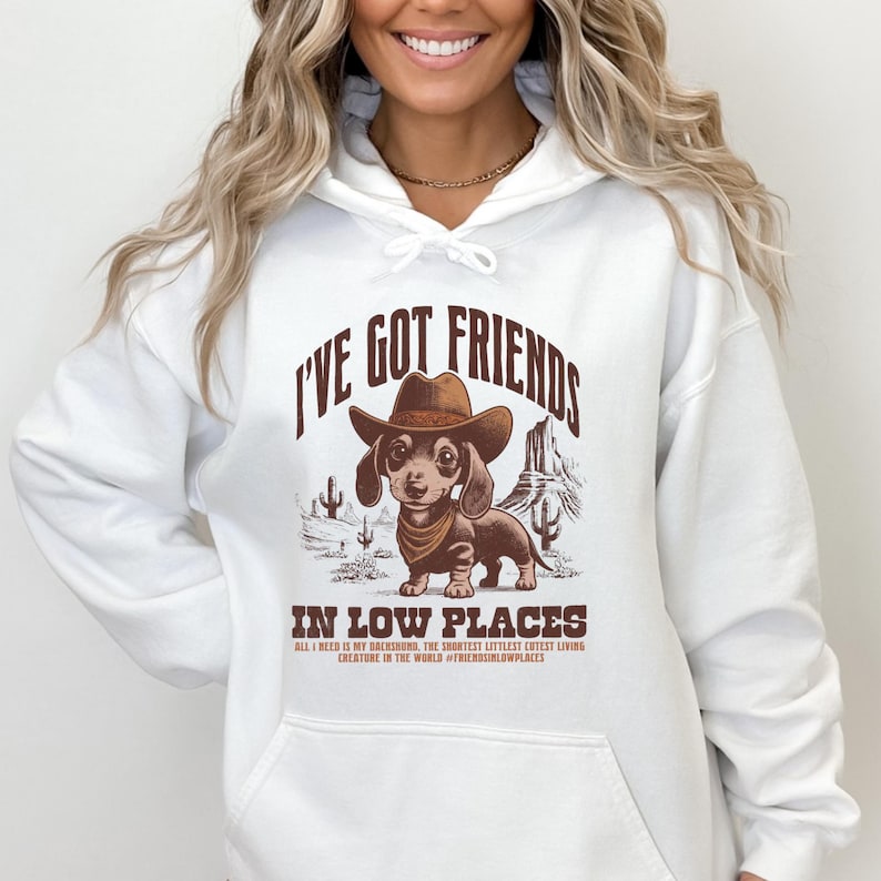 May include: White hoodie featuring a graphic of a dachshund wearing a cowboy hat and bandana, set against a desert landscape. The text reads "I'VE GOT FRIENDS IN LOW PLACES" with additional text below.