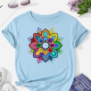 May include: A light blue t-shirt featuring a vibrant, multi-colored mandala design. The mandala is composed of concentric layers of petals in shades of red, orange, yellow, green, blue, and purple. The shirt has short sleeves and a crew neck.