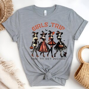 May include: A light gray t-shirt featuring a Halloween-themed graphic. The design includes four women dressed as witches, the words "GIRLS TRIP" and "TIME TO GET WICKED" in orange, and black bats. Each witch holds a pumpkin.