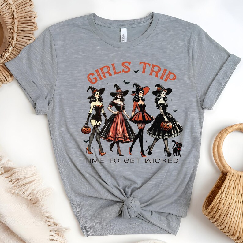 May include: A light gray t-shirt featuring a Halloween-themed graphic. The design includes four women dressed as witches, the words "GIRLS TRIP" and "TIME TO GET WICKED" in orange, and black bats. Each witch holds a pumpkin.
