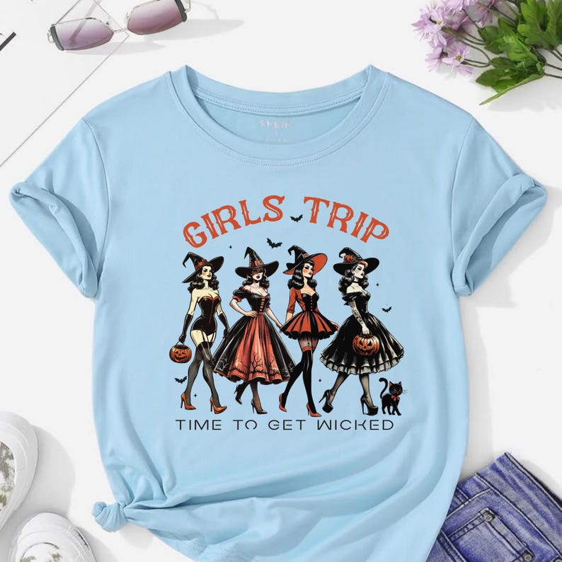 May include: Light blue t-shirt with the words "GIRLS TRIP" and "TIME TO GET WICKED" printed in orange. The shirt features a graphic of four women in witch costumes, holding pumpkins. A black cat is also in the graphic.