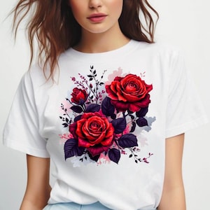 Peut inclure: T-shirt blanc orné d'un motif floral. Le graphisme présente trois grandes roses rouges avec des feuilles et des tiges violet foncé, sur un fond clair et doux. Les roses sont au centre du design.
