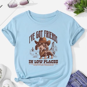 May include: Light blue t-shirt with a graphic of a dachshund wearing a cowboy hat and bandana, with the text "I'VE GOT FRIENDS" and "IN LOW PLACES". The design includes desert scenery and text that reads "ALL I NEED IS MY DACHSHUND, THE SHORTEST LITTLEST CUTEST LIVING CREATURE IN THE WORLD #FRIENDSINLOWPLACES".