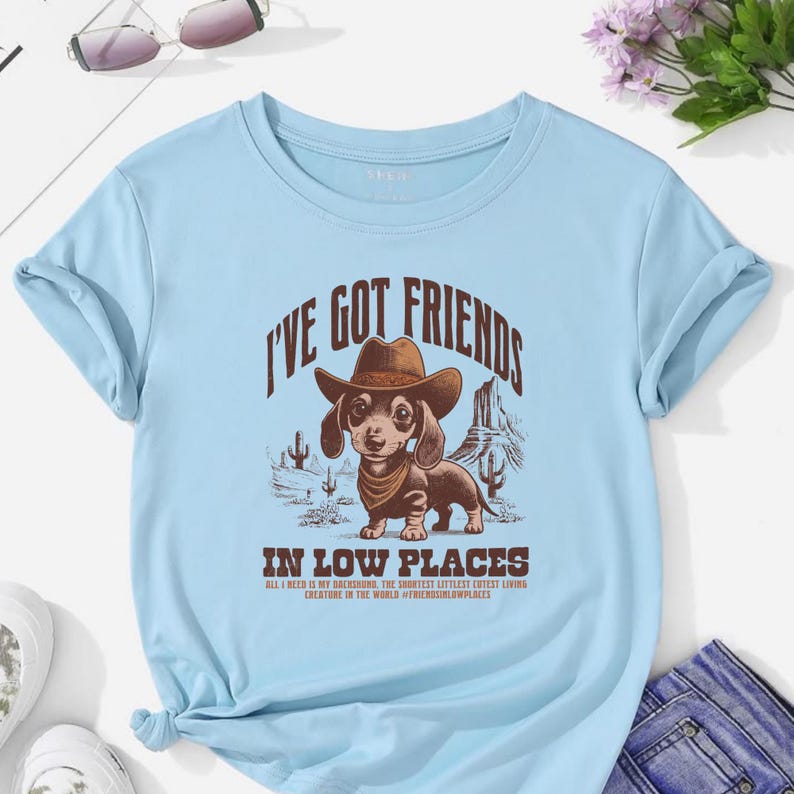 May include: Light blue t-shirt with a graphic of a dachshund wearing a cowboy hat and bandana, with the text "I'VE GOT FRIENDS" and "IN LOW PLACES". The design includes desert scenery and text that reads "ALL I NEED IS MY DACHSHUND, THE SHORTEST LITTLEST CUTEST LIVING CREATURE IN THE WORLD #FRIENDSINLOWPLACES".