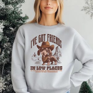 May include: A light gray sweatshirt featuring a dachshund wearing a cowboy hat and bandana. The text "I'VE GOT FRIENDS IN LOW PLACES" is above and below the dog illustration. Additional text reads "ALL I NEED IS MY BACKSHONG, THE SHORTEST LITTLEST CUTEST LIVING CREATURE IN THE WORLD #FRIENDSINLOWPLACES."