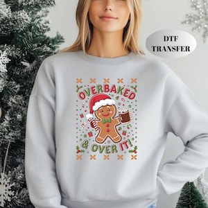 May include: A light gray crewneck sweatshirt featuring a central graphic of a smiling gingerbread person wearing a Santa hat and holding a mug of hot cocoa. The text reads OVERBAKED & OVER IT, surrounded by festive holiday icons like candy canes.