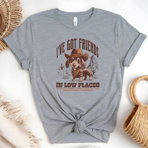 May include: A light grey t-shirt featuring a dachshund wearing a cowboy hat and bandana, with the text "I'VE GOT FRIENDS IN LOW PLACES". The design includes desert scenery. The shirt is knotted at the waist.