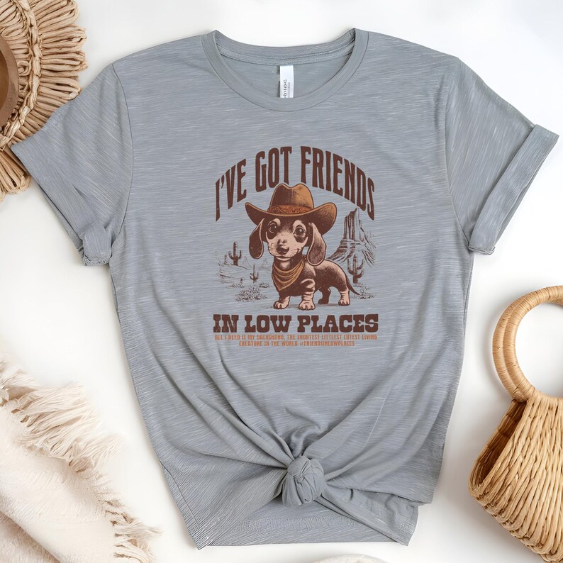 May include: A light grey t-shirt featuring a dachshund wearing a cowboy hat and bandana, with the text "I'VE GOT FRIENDS IN LOW PLACES". The design includes desert scenery. The shirt is knotted at the waist.