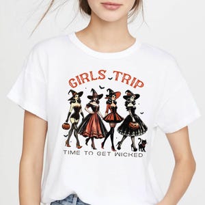 May include: White t-shirt with a Halloween-themed graphic. The design features four women in witch costumes, holding pumpkins, with the words "GIRLS TRIP" and "TIME TO GET WICKED" in orange text.