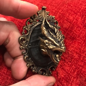 Może przedstawiać: Wisząca dekoracja ścienna w stylu antycznym z głową smoka w kolorze brązu, wyłaniającą się z czarnego owalu. Smok ma szczegółowe łuski i otwartą paszczę. Owal jest obramowany ozdobną, brązową obwódką.