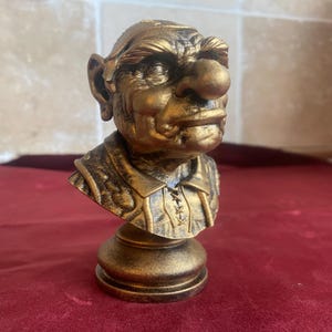 Op de afbeelding: Een bronzen buste van een trol met een grote neus, opvallende oren en een strenge uitdrukking. De trol draagt een gedetailleerde tuniek met een veterlijn. De buste is gemonteerd op een ronde basis, waardoor een decoratieve sculptuur ontstaat.