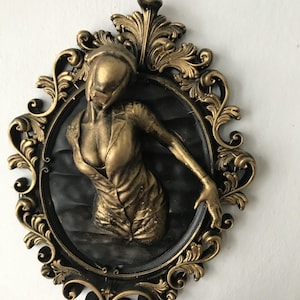 Puede incluir: Escultura en bajorrelieve dorada de una figura en un marco de estilo vintage. La figura lleva un vestido o uniforme, con un fondo negro ovalado. El marco es ornamentado con un acabado dorado oscuro.