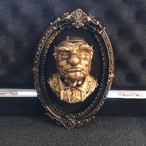 Op de afbeelding: Ovale wandplaat in antieke stijl met een goudkleurige buste van een man met een strenge blik, een hoed en gedetailleerde kleding. De buste staat tegen een zwarte achtergrond in een sierlijk frame met een bronzen afwerking.