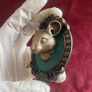 May include: A bronze-colored mouse head sculpture set against a green background, framed by an ornate, dark bronze border. The sculpture is held by a gloved hand, with a red background.