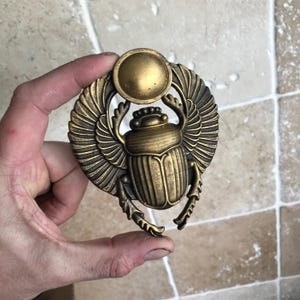 Könnte beinhalten: Ein bronzefarbener Skarabäus-Anhänger mit ausgebreiteten Flügeln und einer Sonnenscheibe. Der Anhänger, gehalten in einer Hand, weist aufwendige Details auf, die an altägyptische Kunst erinnern. Der Käfer symbolisiert Wiedergeburt.
