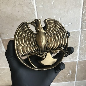 Puede incluir: Una escultura de metal de color dorado de un pájaro con las alas extendidas. El pájaro está posado en una rama y está rodeado de un diseño circular.