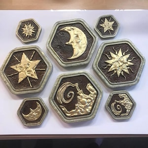 Puede incluir: Ocho azulejos hexagonales de cerámica con detalles dorados que representan símbolos celestiales. Los símbolos incluyen una luna creciente, un sol con cara, una estrella y una imagen estilizada de un elemento de agua.
