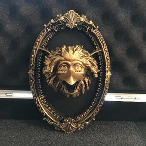 Puede incluir: Adorno de pared ovalado y ornamentado, con una criatura dorada con forma de pájaro con pico abierto, sobre un fondo negro. El marco es de color oro oscuro con detalles intrincados.
