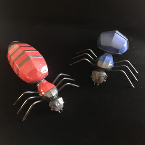 May include: Two metal spiders, one red and one blue, with black legs. The spiders are made of metal and have a geometric design.