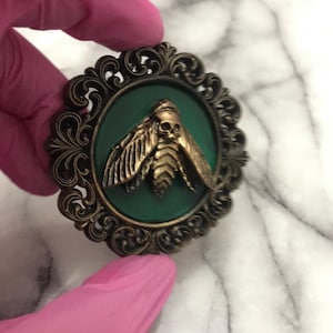 May include: A bronze moth brooch with a skull-shaped head and detailed wings, set against a dark green background. The moth is framed by an ornate, antique-style bronze border. A unique, gothic-inspired accessory.