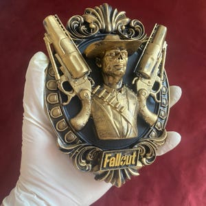 Könnte beinhalten: Goldfarbene Wandplakette mit einer Büste eines Mannes mit Hut, flankiert von zwei Revolvern. Die Plakette hat einen schwarzen Hintergrund und das Wort "Fallout" unten. Die Plakette wird von einer behandschuhten Hand gehalten.